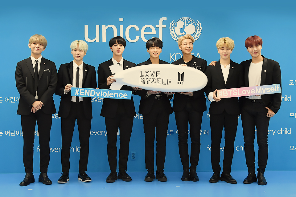 A photo of BTS standing in a line in front of a blue wall that says unicef. The members all wear black suits and white dress shirts. They hold signs that say #ENDviolence, #BTSLoveMyself, and a model blimp that has the words Love Myself and the BTS logo on the side