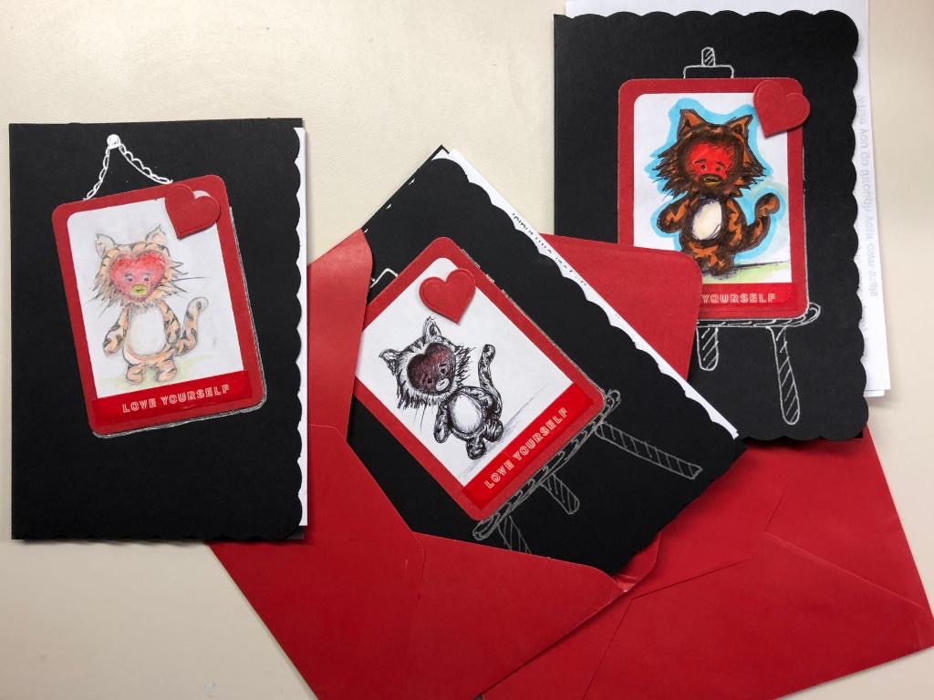 Photo of 3 pieces of hand drawn art. Each picture is of a cartoon tiger standing on its hind legs. They all have BT21 character Tata's face. Each drawing is mounted on a piece of black cardstock. Two pieces of cardstock have a drawing of an easel on them, making it look like the tiger drawings were painted using the easels. The third cardstock has a drawing of a nail and twine, making it look like the tiger drawing is hanging on a wall. There are red cardstock hearts in the top right corner of each drawing. Underneath each drawing are the words love yourself
