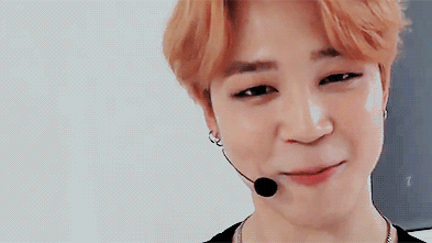 Gif of Jimin smiling and waving at the camera. Jimin has light orange hair styled in a bowl cut with a center part in the bangs. His eyes are heavily lined in black and he wears a small, hands-free mic over his right ear.