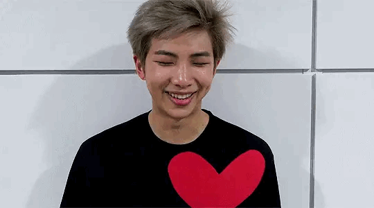 Gif of Namjoon with an open mouth smile. He is visible from the chest up and wears a black t-shirt. His eyes are closed and they tighten the wider he smiles. He has short, ash blond hair that is slightly spiked on top and shorter on the sides of his head. He holds a red, cardboard heart in front of him.