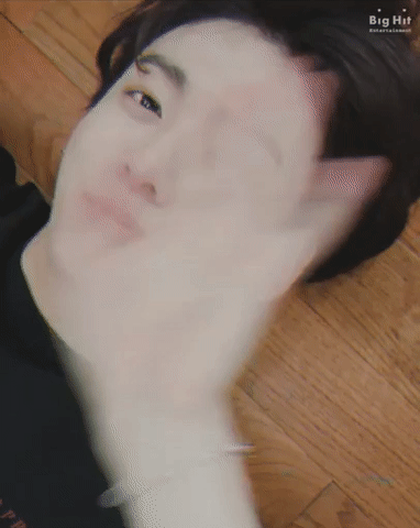 A gif of Jungkook lying on a wooden floor, waving at the camera. He's wearing a black t-shirt and has a ring piercing on his right eyebrow.