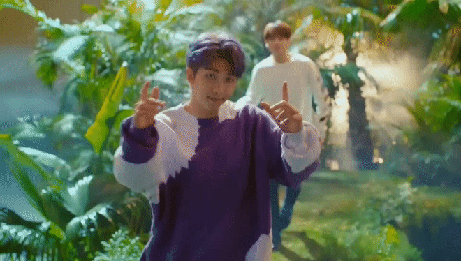 Gif of Namjoon making finger guns and winking at the camera. Namjoon is walking through a tropical forest. He wears a purple and white long-sleeved shirt and has purple hair, styled with a side part.