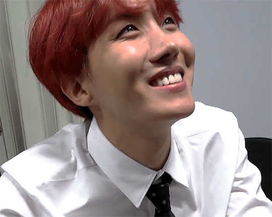 Gif of Hoseok smiling towards the top right of the frame. He's wearing a white button-up shirt with a black tie and has dark red hair styled in a bowl cut with a center split in the bangs.