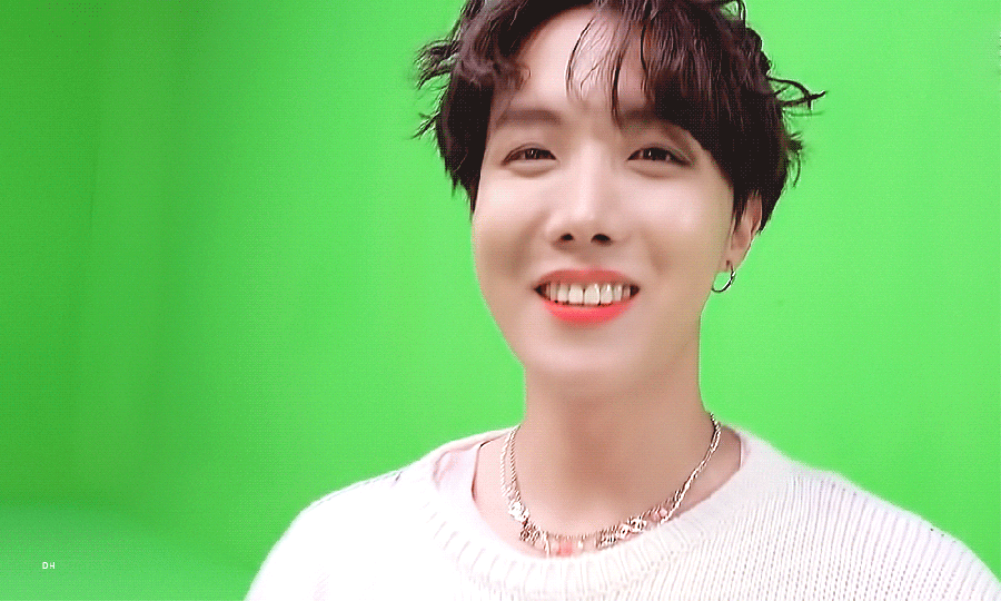 Gif of Hoseok holding up his nails to the camera. With his right hand he makes the rock on sign, showing that his pointer finger nail is painted black with the word "Hope" in white and a yellow smiley face on his pinky finger. With his left hand he holds up his pointer, middle, and ring finger and shows that there's one letter on each nail, spelling Ego. Hoseok wears a white knit sweater and has brown hair styled in a bowl cut with a center part in bangs.