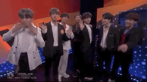 Gif of BTS in a television studio waving at the camera. All members are wearing suits with white, black, or grey jackets. Their suit jackets are open to reveal white, red, or grey button up shirts.