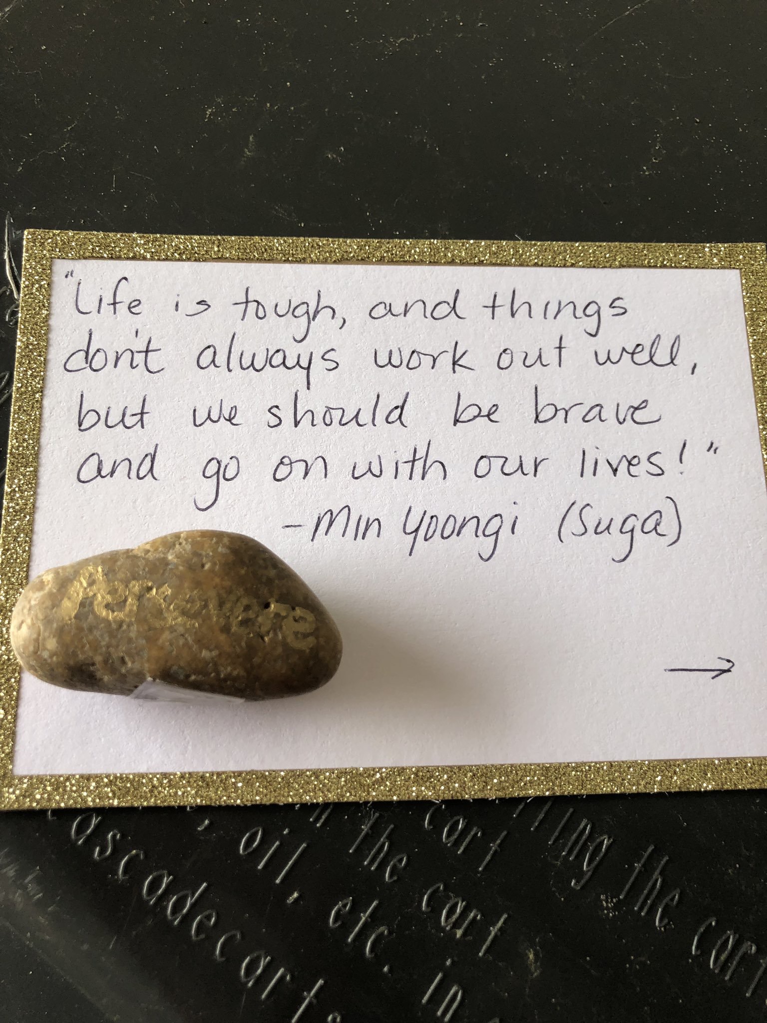 A photo of a rock with a note that contains a quote from Min Yoongi or Suga. It says "life is tough, and things don't always work out well, but we should be brave and go on with our lives!"