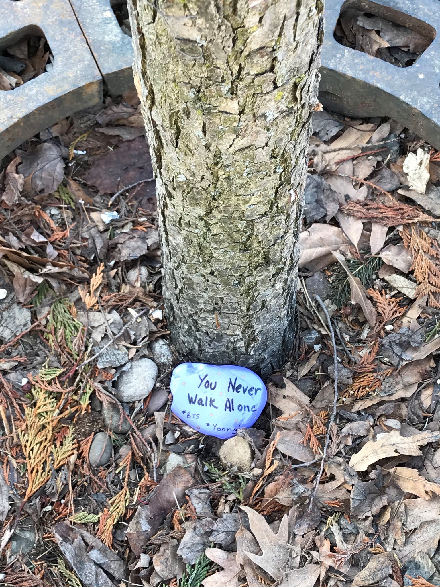 A photo of a rock painted purple resting against the base of a medium sized tree. The words "you never walk alone #BTS #Yoongi" are painted on the stone