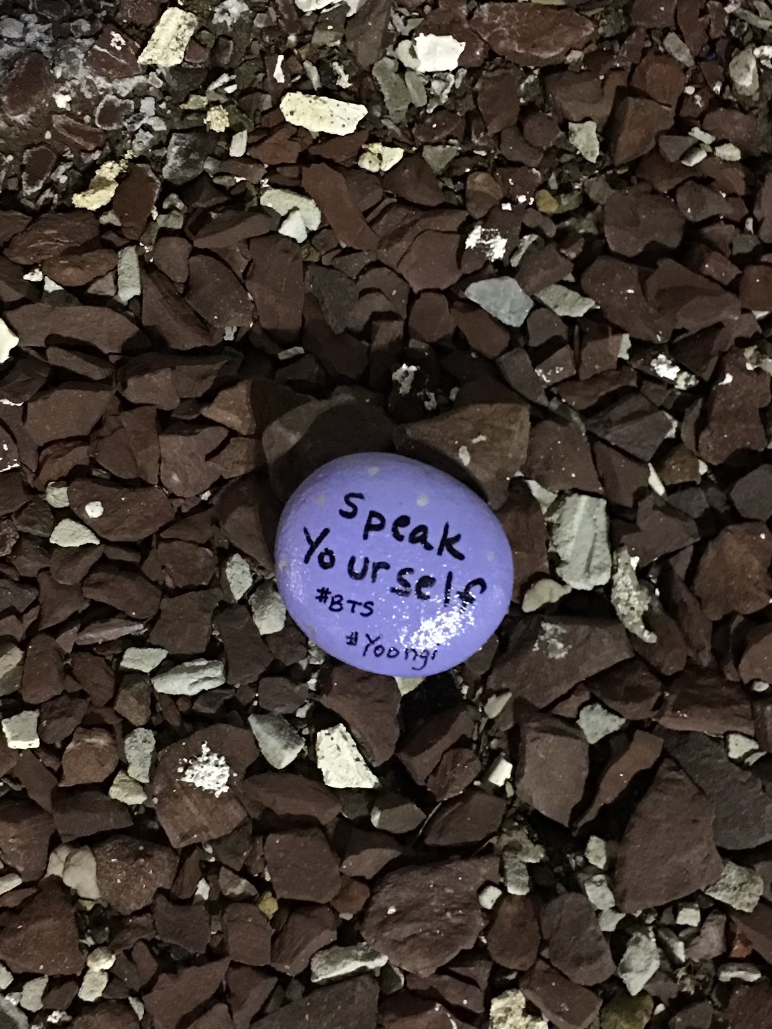 A photo of a rock painted purple placed on top of dark brown gravel. The words "speak yourself #BTS #Yoongi" are painted on the stone