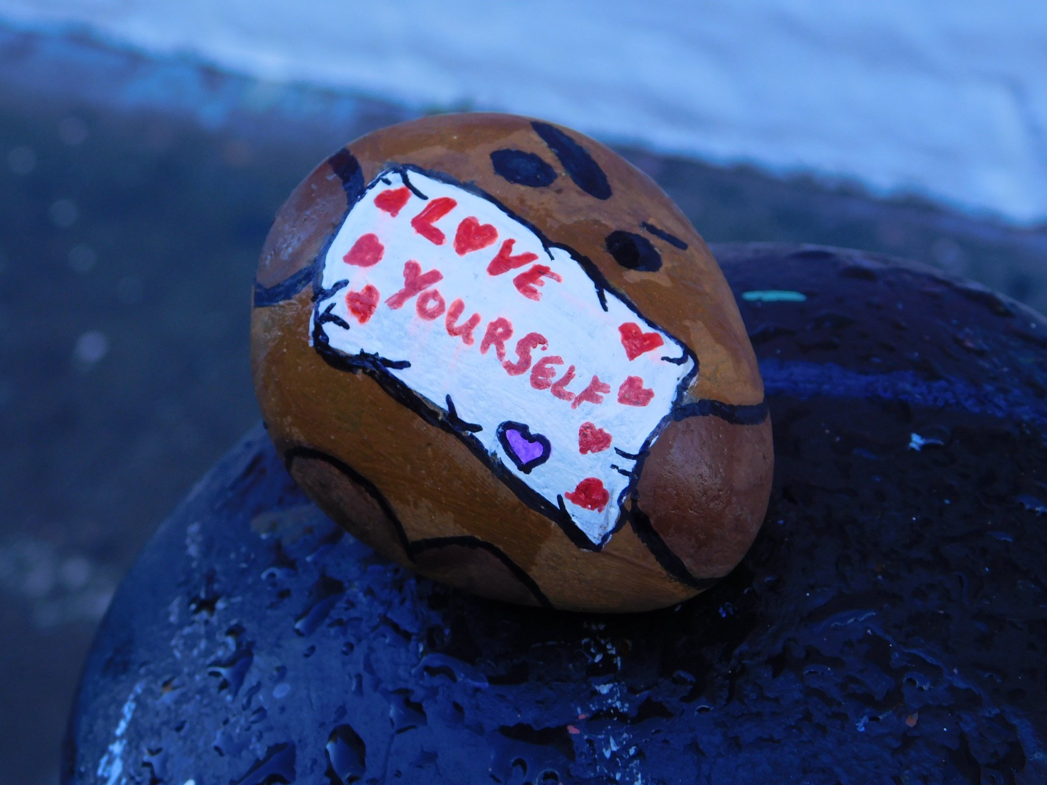 A photo of a rock painted to look like BT21 character Shooky holding a sign that says "love yourself"