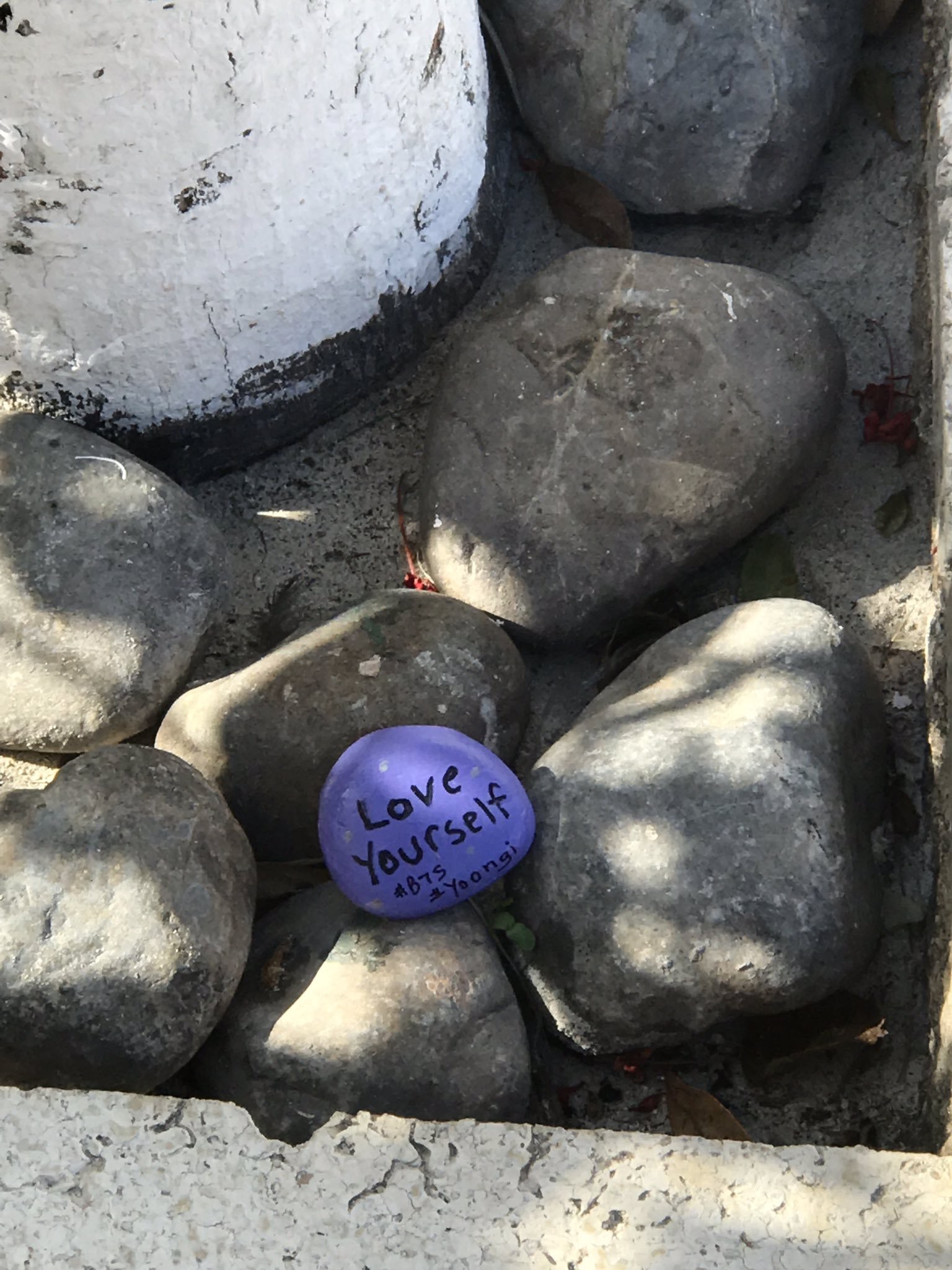 A photo of a painted purple rock nestled among larger rocks. The rock has the words "love yourself #BTS #Yoongi" on it