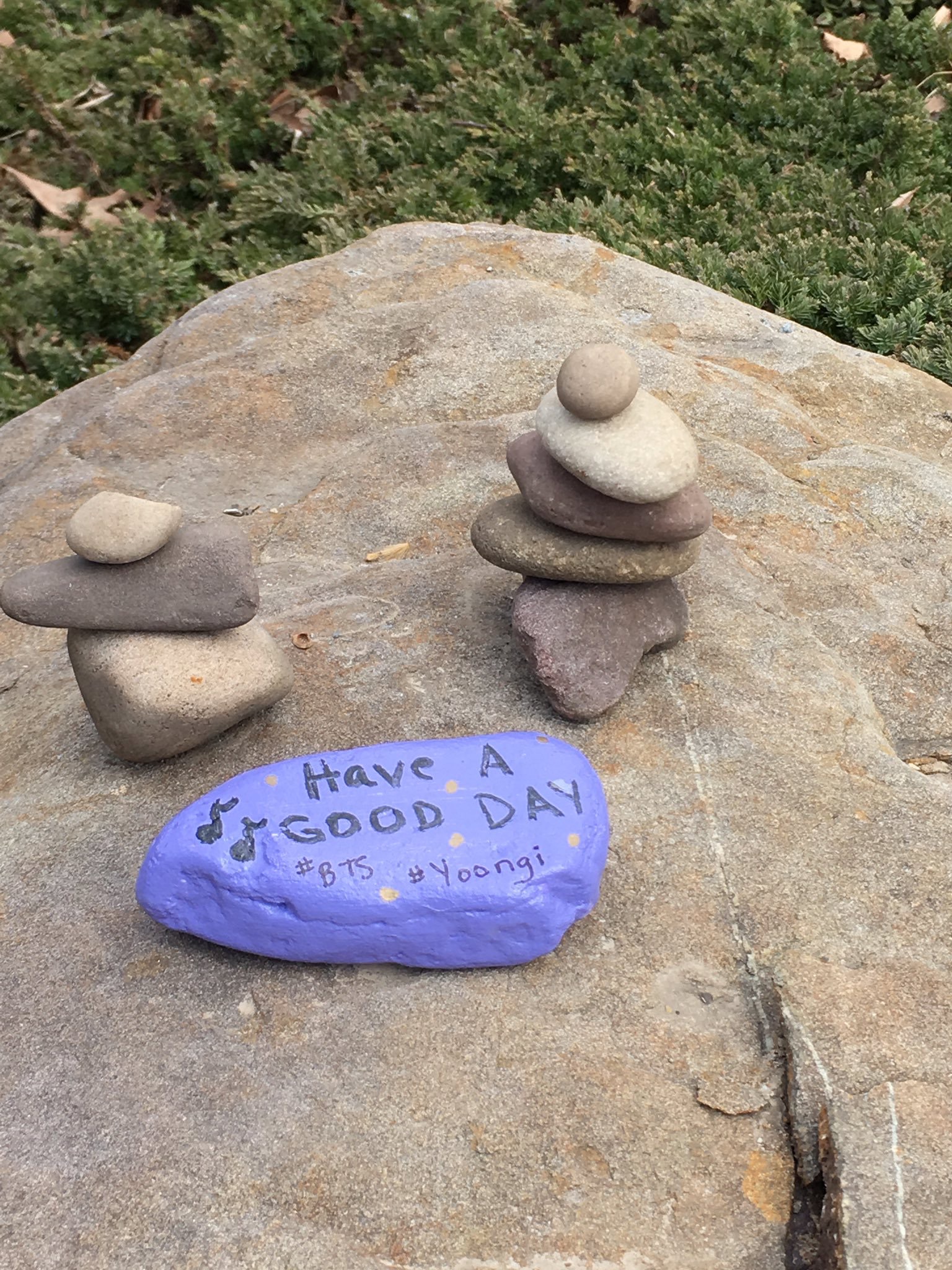 A photo of a rock painted purple with the message "have a good day #BTS #Yoongi". There are two small stacks of stones behind the purple rock.