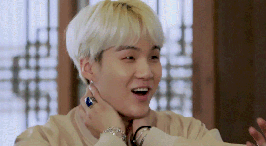 A gif of Yoongi laughing. He's wearing an off-white jacket and is pressing both hands to his neck in surprise. His hair is platinum blond and styled in a bowl cut with a slight center part to the bangs